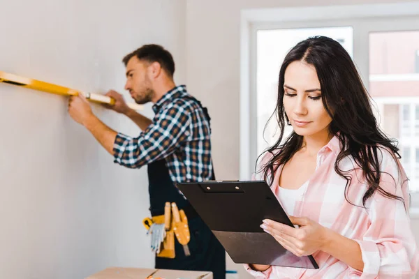 Selective focus of attractive woman holding clipboard with handyman measuring wall on background — Stock Photo