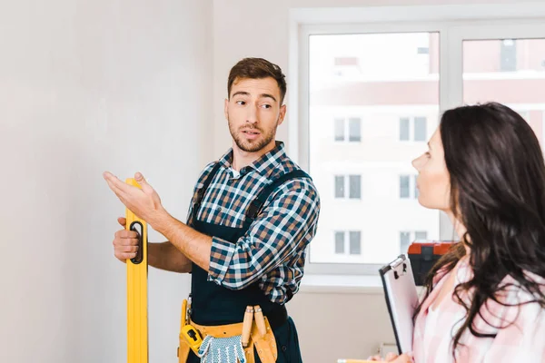 Woman holding clipboard and looking at wall while handyman gesturing and holding measuring level — Stock Photo
