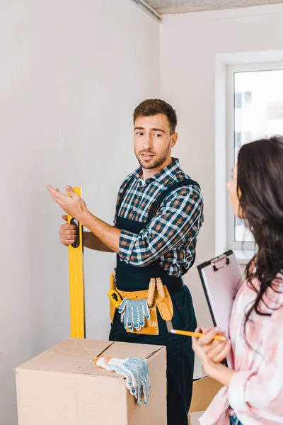 Selektiver Fokus des Handarbeiters, der mit Klemmbrett in der Nähe einer Frau gestikuliert und den Messpegel hält und auf die Wand blickt — Stockfoto