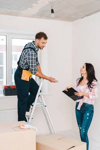 Alegre mujer sosteniendo portapapeles y mirando al manitas de pie en la escalera - foto de stock