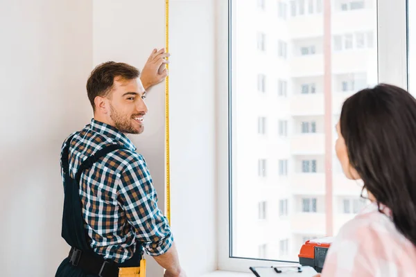 Selective focus of cheerful handyman measuring window near female client — Stock Photo