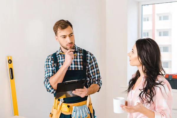 Thoughtful handyman holding clipboard and looking at attractive woman with cup — Stock Photo