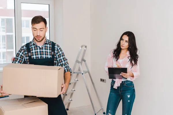 Attractive woman holding clipboard and looking at handsome handyman with box in hands — Stock Photo