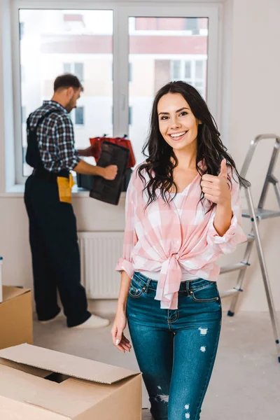 Selective focus of cheerful woman showing thumb up with handyman on background — Stock Photo