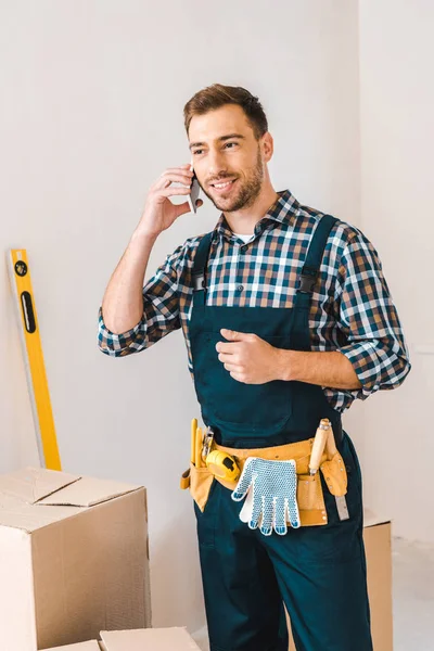 Handsome handyman talking on smartphone while standing with tool belt on waist — Stock Photo