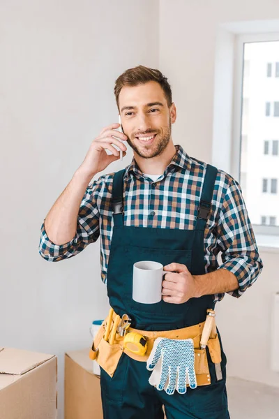 Handsome handyman talking on smartphone while standing with cup of drink — Stock Photo
