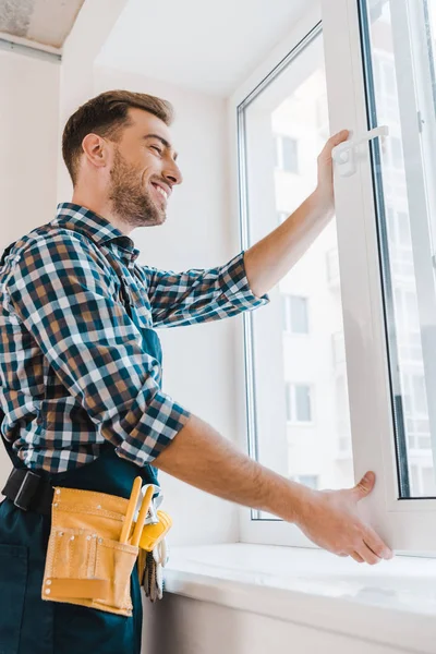Cheerful handyman with tool belt smiling while opening window — Stock Photo