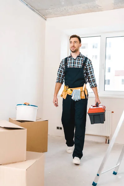 Guapo manitas sosteniendo caja de herramientas y caminando cerca de cajas en la habitación - foto de stock