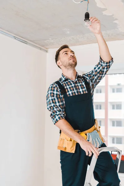 Handsome handyman standing on ladder and looking at light bulb — Stock Photo