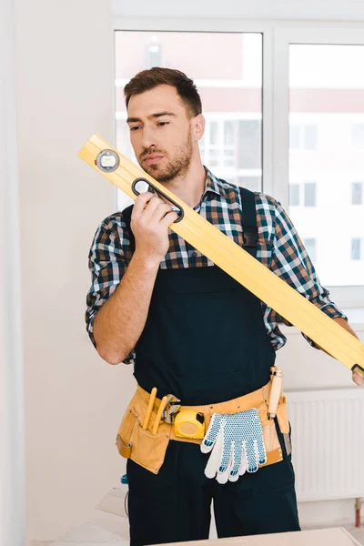 Handyman bonito olhando para o nível de medição enquanto está em pé no quarto — Fotografia de Stock