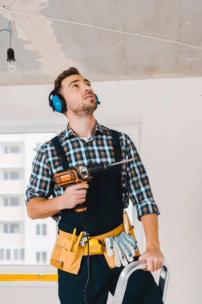 Handsome handyman holding drill in hand and looking on ceiling — Stock Photo