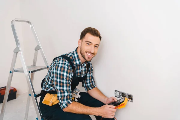 Cheerful handyman holding digital multimeter and sitting near power socket — Stock Photo