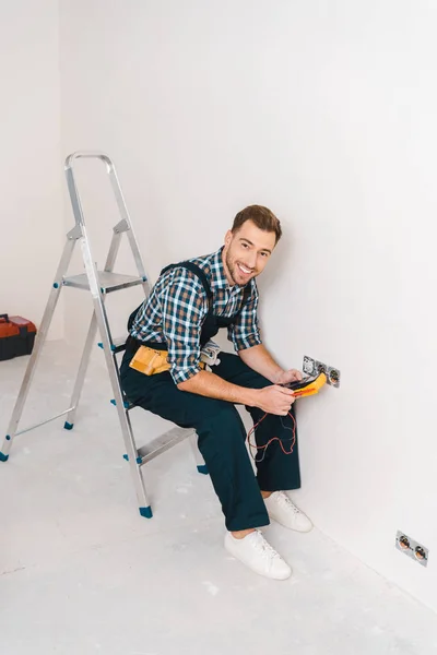 Happy handyman holding digital multimeter and sitting on ladder near power socket — Stock Photo