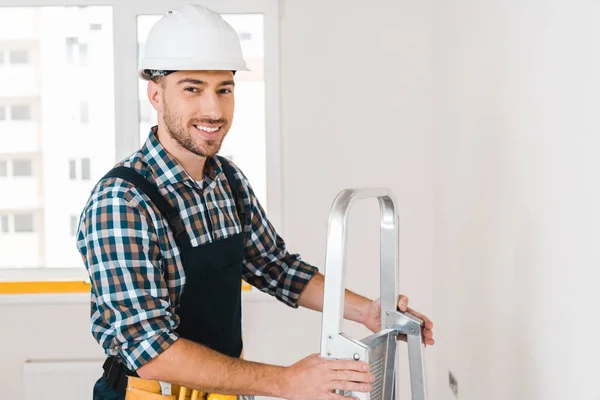Cheerful handyman in helmet smiling while holding ladder — Stock Photo