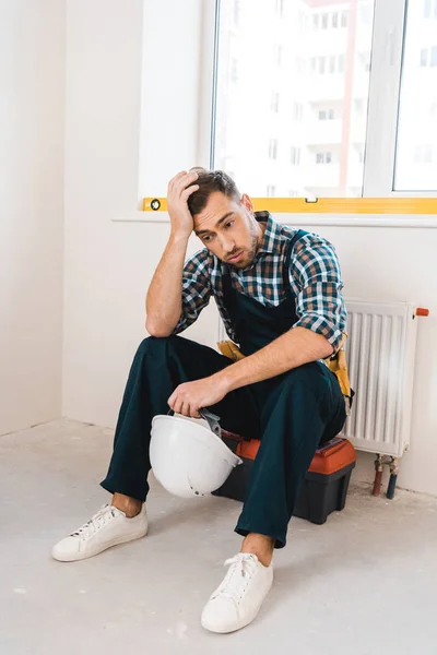 Erschöpfter Handwerker sitzt auf Werkzeugkiste und hält Helm — Stockfoto