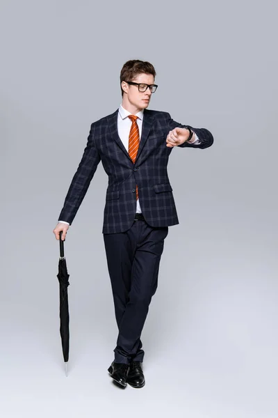 Elegante joven hombre de negocios con paraguas mirando reloj de pulsera aislado en gris - foto de stock