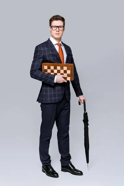 Молодой бизнесмен с шахматной доской и зонтиком изолированы на сером — стоковое фото