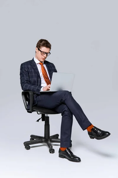 Uomo d'affari seduto sulla sedia e al lavoro sul computer portatile su grigio — Foto stock