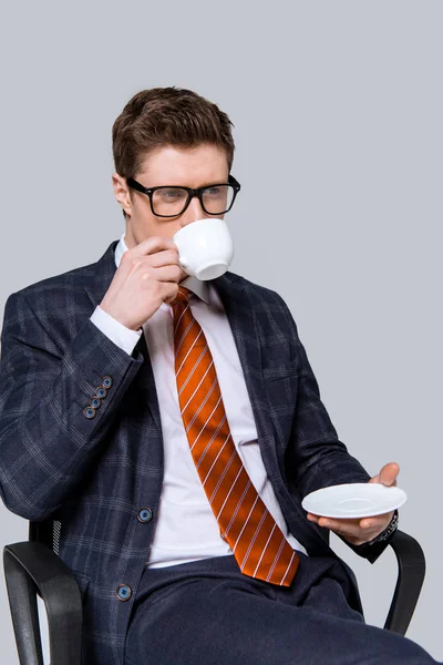 Handsome stylish businessman drinking coffee on chair isolated on grey — Stock Photo