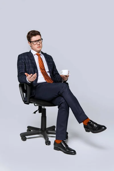 Stylish businessman sitting on chair with cup of coffee on grey — Stock Photo