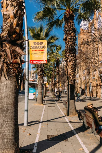 BARCELONA, SPAGNA - 28 DICEMBRE 2018: strada cittadina con alte palme verdi e persone sedute su panchine — Foto stock