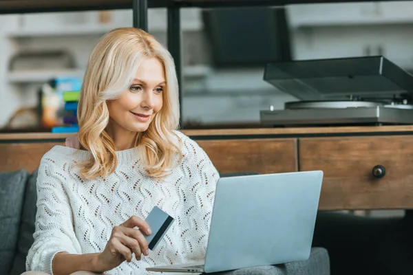 Mujer alegre que sostiene la tarjeta de crédito mientras que usa el ordenador portátil en casa - foto de stock