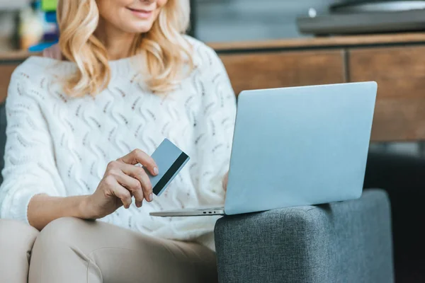 Vista recortada de la mujer alegre que sostiene la tarjeta de crédito mientras usa el ordenador portátil en casa - foto de stock