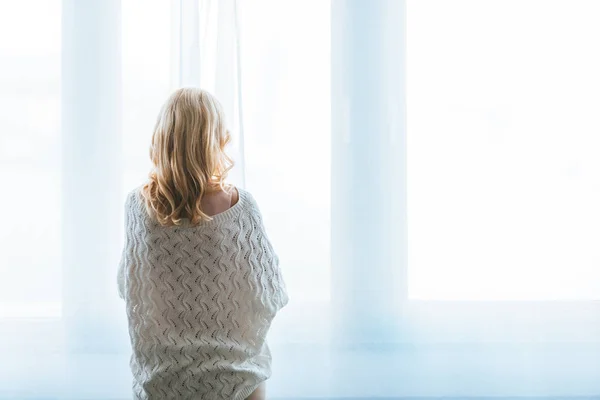 Rückansicht einer Frau mit blonden Haaren, die zu Hause am Fenster steht — Stockfoto