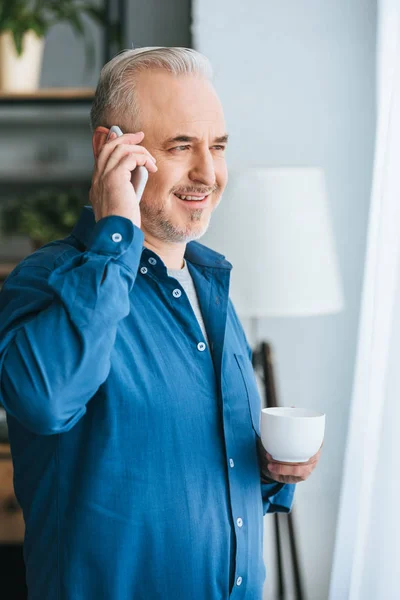 Hombre alegre sosteniendo la taza y sonriendo mientras habla en el teléfono inteligente - foto de stock