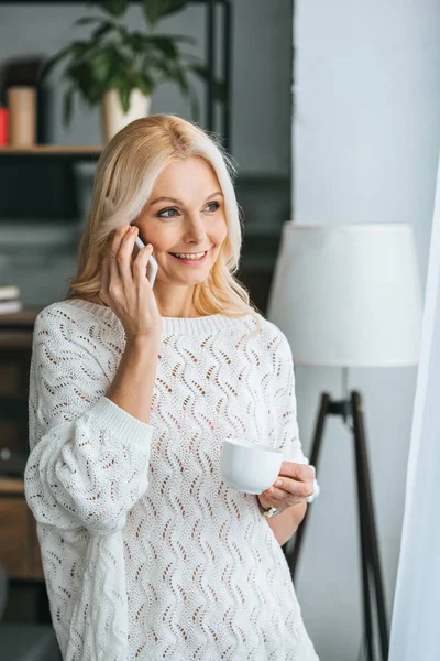 Mujer rubia sosteniendo la taza y sonriendo mientras habla en el teléfono inteligente - foto de stock