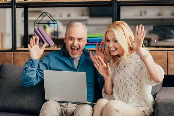 Feliz pareja mirando a la computadora portátil y saludando las manos mientras tiene videollamada - foto de stock