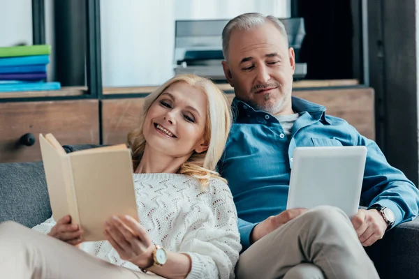 Fröhliche Frau liest Buch neben glücklichem Ehemann mit digitalem Tablet, während sie auf dem Sofa sitzt — Stockfoto