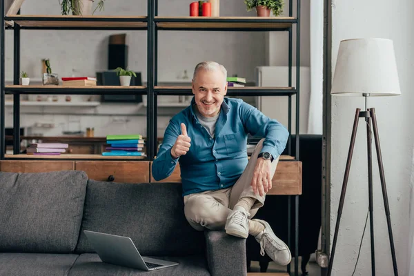 Щасливий чоловік показує великий палець під час сидіння на дивані біля ноутбука — стокове фото