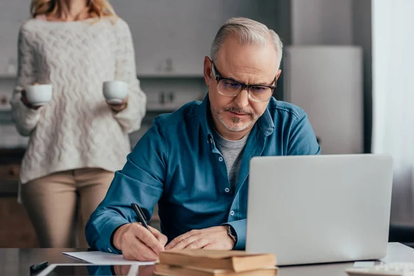 Selective focus of concentrated man in glasses writing near laptop with wife holding cups on background — Stock Photo