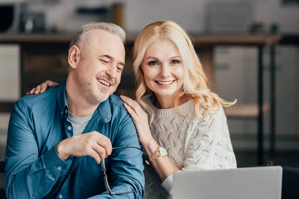 Homem bonito segurando óculos e sorrindo com esposa alegre enquanto olha para laptop — Fotografia de Stock
