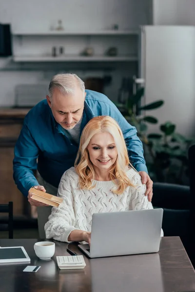 Handsome man holding books near smiling wife using laptop — Stock Photo