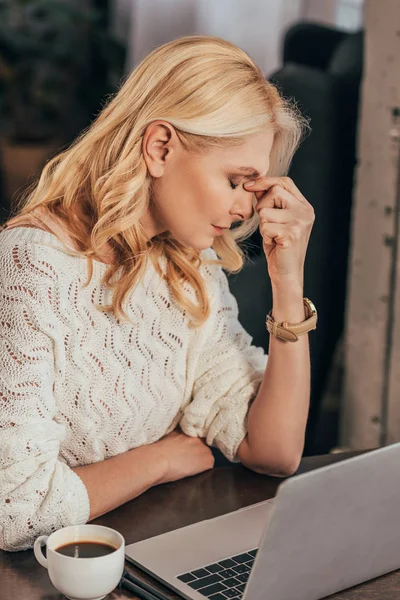 Mulher cansada sentada com os olhos fechados perto de laptop e xícara com café — Fotografia de Stock