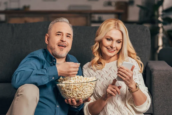 Felice uomo che tiene ciotola con popcorn vicino moglie utilizzando smartphone — Foto stock