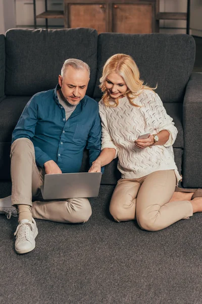 Счастливая женщина держит смартфон и смотрит на ноутбук рядом с мужем дома — стоковое фото