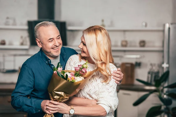 Cheerful man smiling while giving flowers to attractive wife at home — Stock Photo