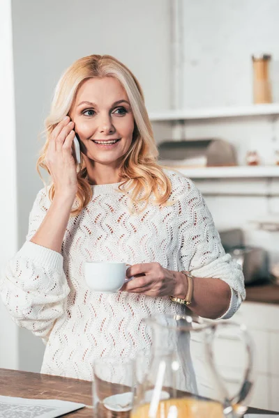 Cheerful woman talking on smartphone while holding cup with drink in kitchen — Stock Photo