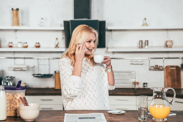 Cheerful woman talking on smartphone while drinking coffee in kitchen — Stock Photo