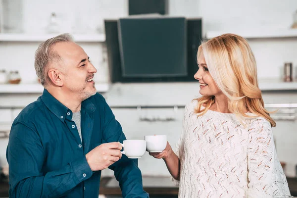 Glückliche Frau hält Tasse in der Hand und schaut fröhlichen Mann zu Hause an — Stockfoto