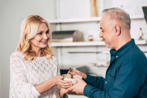 Atractivo esposa celebración bowl con cornflakes cerca alegre marido en cocina - foto de stock