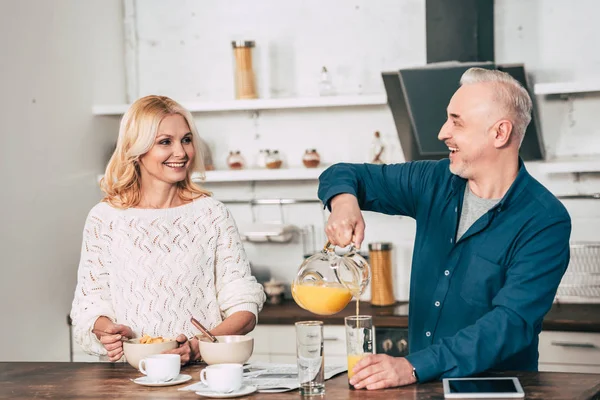 Fröhlicher Mann gießt Orangensaft in Glas neben attraktiver Frau — Stockfoto