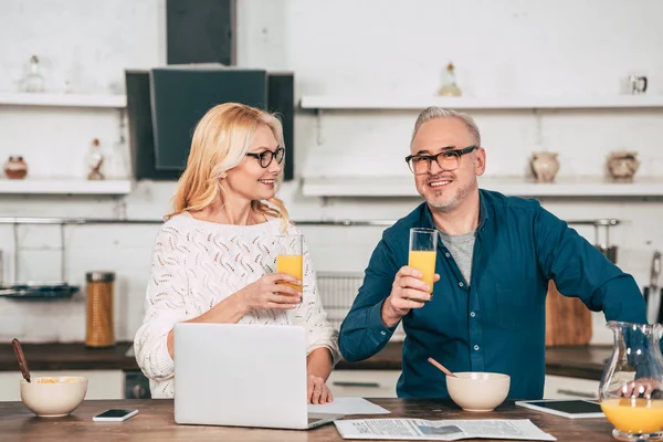 Femme gaie regardant mari dans des lunettes de vue tenant verre avec du jus d'orange près d'un ordinateur portable — Photo de stock