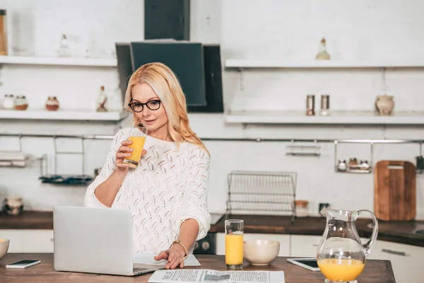 Attraktive Frau in Brille trinkt Orangensaft in der Nähe von Laptop — Stockfoto