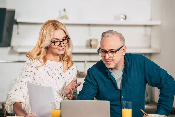 Attraktive Frau hält leeres Papier in der Hand und steht mit Mann in Brillennähe neben Laptop — Stockfoto