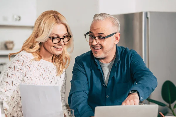 Happy woman holding blank paper and standing with husband in eye glasses near laptop — Stock Photo