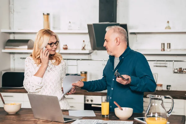 Atractiva mujer hablando en el teléfono inteligente y de pie con el marido en la cocina cerca de la computadora portátil - foto de stock